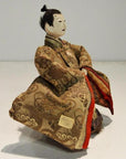 White-faced Japanese Taisho samurai doll in ornate robe, seated with folded arms, close-up view.