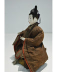 Brown fabric samurai doll with black hair ornament, seated with hands raised, indoors.