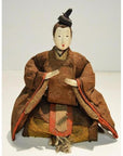 Close-up of Japanese Taisho Samurai doll in brown kimono, seated with folded hands and small hat, fabric details visible.