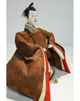 Close-up of Japanese Taisho era Samurai doll in brown kimono with red lining, seated against plain background.