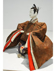 Close-up of Japanese Taisho era samurai doll with painted face, patterned brown kimono, red and white fabric accents.