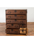 Dark wood Japanese Taishō Tansu chest with six drawers and a safe, on wooden floor, white brick wall background.