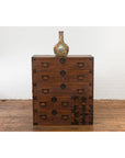 Dark wood Japanese Taishō Tansu chest with six drawers and safe, vase on top, against white brick wall background.