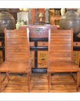 Brown teak wood side chairs beside each other in front of a vintage cabinet with painted floral details and vases indoors.