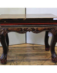 Black lacquered wooden side table with carved guardian design and worn finish, on wooden floor against plain backdrop.