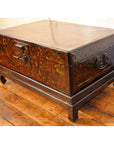 Black lacquered wood trunk with floral designs, metal handles, and lock on stand, placed on a wooden floor.