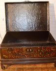 Black lacquered trunk with ornate floral designs on stand, shown on a wooden floor indoors.