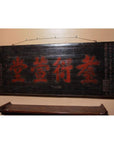 Red Chinese characters on 19th century wooden signboard, mounted above a wooden shelf on a beige wall.