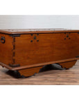 Brown teak wood chest with iron nailheads and wheels on a wooden floor next to a white brick wall indoors.