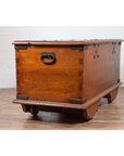 Brown teak wood blanket chest with iron nailheads on wheels, placed on a wooden floor by a white wall indoors.