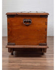 Brown teak wood chest with iron nailheads and wheels on wooden floor against a white brick wall indoors.