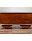 Brown teak wood chest with iron nailheads and wheels on a wooden floor, positioned against a white wall indoors.