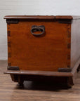 Brown teak wood chest with iron nailheads and wheels on a wooden floor against a white brick wall indoors.