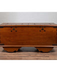 Brown teak wood chest with iron nailheads and wheels on wooden floor next to a white brick wall indoors.