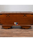 Brown teak wood blanket chest with iron nailheads on wheels, placed on wood floor next to a white brick wall indoors.