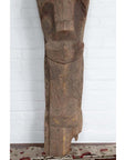 Brown hand-carved wooden Singa Singa tribal carving from Batak, Sumatra, against a white brick wall on patterned carpet.