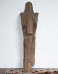 Brown wood Singa Singa tribal carving from the Batak people of Sumatra, on patterned cloth with brick wall background.