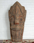Brown wood Singa Singa tribal mask from Batak people, Sumatra, on patterned cloth against a white brick wall.
