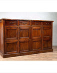 Brown teak wood cabinet with four drawers and four double doors, against white brick wall on wood floor indoors.