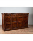Brown teak wood cabinet with four drawers and four double doors, set against a white brick wall indoors.