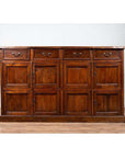 Brown teak wood cabinet with four drawers and four double doors, set against a white brick wall indoors.