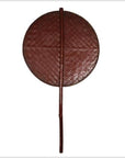 Close-up of a round, reddish-brown woven fan with a long handle, made from natural fibers.