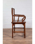 Brown wooden wedding chair with carved medallion and curving arms, side view, indoors against white brick wall.