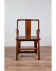 Brown wooden wedding chair with carved medallion and curving arms, placed next to a white brick wall indoors.