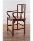 Brown wooden wedding chair with carved medallion and curving arms placed indoors by a white brick wall.