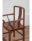 Brown wooden Ming Dynasty style wedding chair with carved medallion and curving arms next to white brick wall indoors.