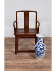 Brown wooden Ming Dynasty style wedding chair next to blue and white floral ceramic vase on wooden floor indoors.