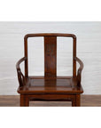 Brown wooden chair with curving arms and carved medallion, placed indoors against a white brick wall.