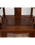 Close-up of brown wooden Ming Dynasty style wedding chair with carved medallion and curved arms on wood floor.