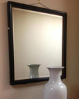 Large white vase reflected in a mirror, with lighter wood-accented wall in the background.