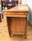 Natural wood altar coffer with scalloped edges and brass hardware, placed near shelves of colorful vases.