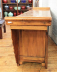 Natural wood altar coffer with scalloped edges and brass hardware, placed near shelves of colorful vases.