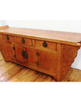 Natural wood altar coffer with ornate carvings and brass hardware, displayed on a wooden floor.