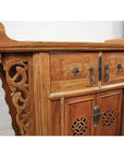 Natural elmwood altar cabinet with floral carvings, ornate details, and metal handles against a white brick wall.