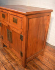 Natural wood small cabinet with two drawers, unique hardware, and brass handles in front of a white brick wall.