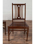Brown wood Indonesian side chairs with pierced backrests, on wooden floor near a white brick wall indoors.