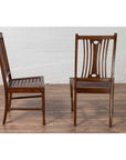 Brown wood Indonesian side chairs with pierced splats on wood floor, white brick wall background, indoor setting.