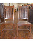 Pair of teak armless chairs on wooden floor in antique shop, side by side, with antique furnishings in background.