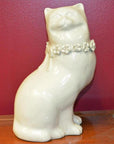 White porcelain cat figurine with floral collar, head turned to side, on wooden surface against red wall.