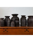 Brown rustic wooden ghee pots in groups of 3, 5, and 7 against a white brick wall indoors.
