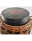 Black rattan and bamboo lidded basket with woven sides, handles, and red floral lid design, close-up view.