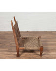 Brown wooden low chair with rope seat, rustic and weathered, on wooden floor by a white brick wall indoors.
