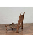 Brown wooden low chair with rope seat and weathered finish on a wooden floor next to a white brick wall indoors.