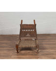 Brown wood and rope low Indian chair with weathered finish on wooden floor near a white wall indoors.
