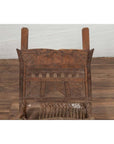 Brown wooden low chair with rope seat, weathered finish, placed on a wooden floor beside a white brick wall indoors.