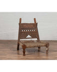 Brown wooden low chair with rope seat, weathered look, on wooden floor next to white brick wall indoors.